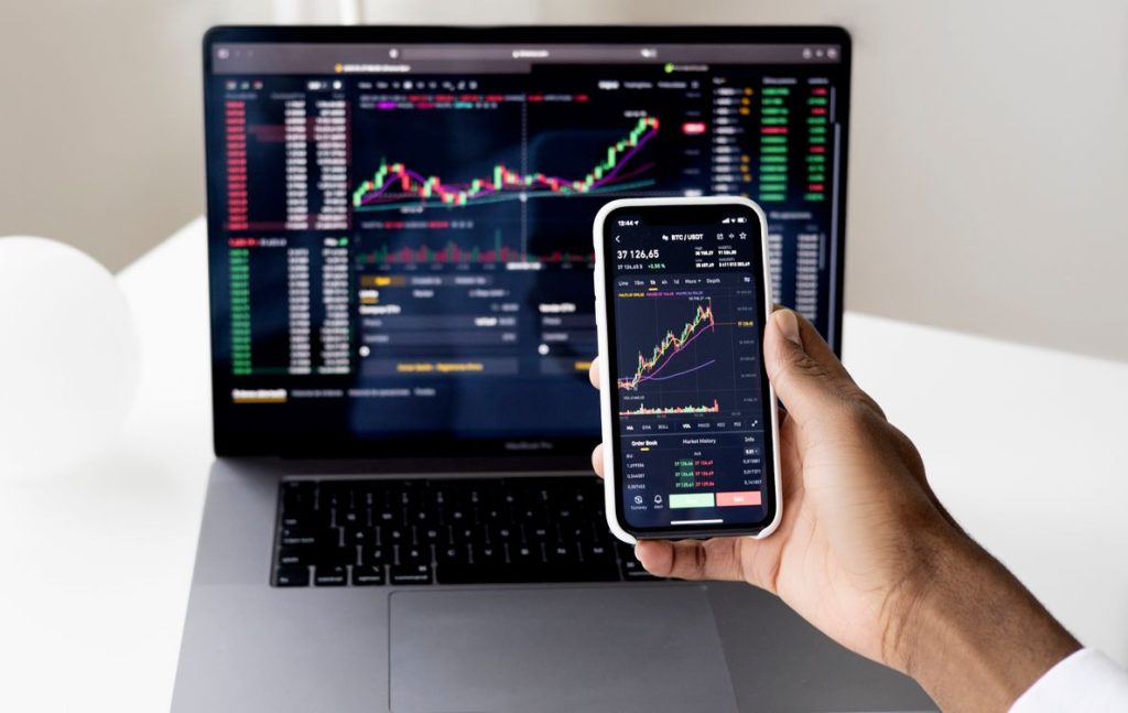 Hand holding a smartphone showing trading charts in front of a laptop